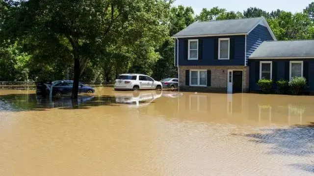Brown floodwater surrounding a two-story home and parked cars after heavy storm rain